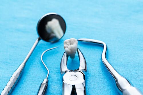 Dental tools including a mirror, pliers holding a tooth, and pick-like instruments on a blue surface.
