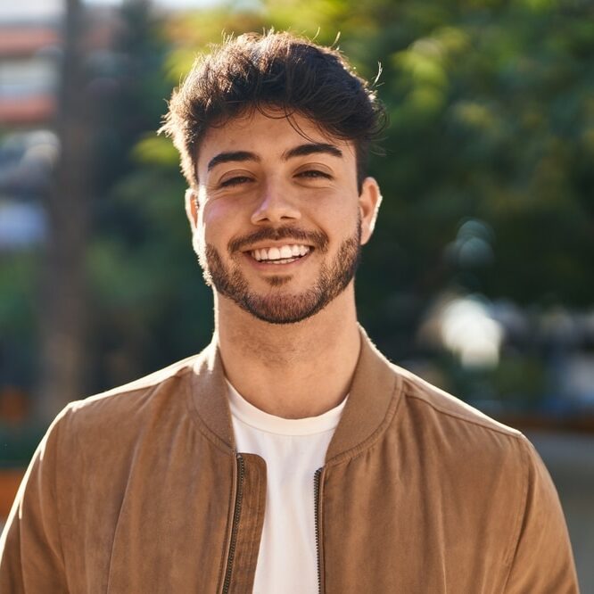 A young man smiling in casual clothes while standing outside in front of an apartment building and some trees.